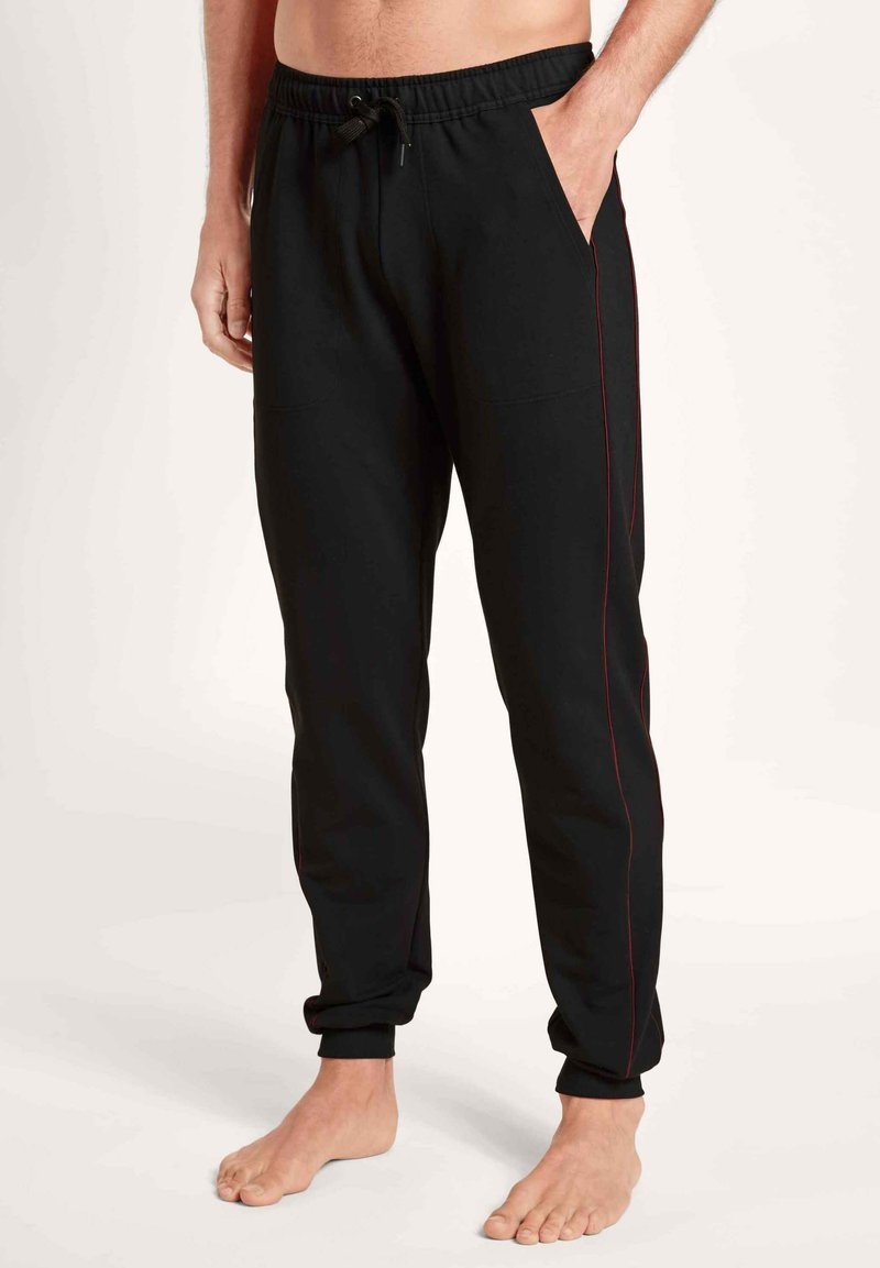 Pantalons de jogging noirs avec une bande latérale rouge, taille élastique et poches latérales. Portés par une personne pieds nus. Design décontracté.