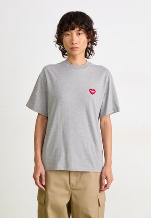 Personne avec des cheveux courts et bouclés, portant des boucles d'oreilles en argent, un t-shirt gris avec un patch en forme de cœur rouge et un pantalon cargo beige devant un fond uni.