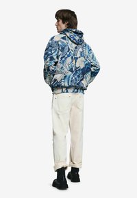 Modrá mikina s kapucňou s prepracovanými vzormi, v kombinácii s bielymi ošúchanými džínsami a čiernymi členkovými čižmami. Mäkká látka, voľný strih.