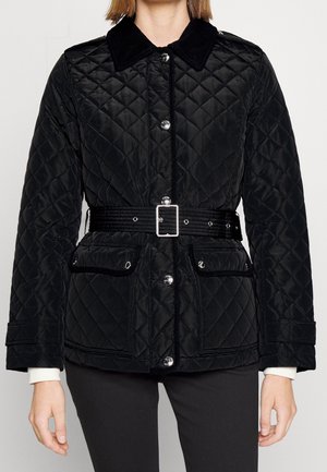 Chaqueta negra acolchada con cinturón, botones plateados, bolsillos con solapa y cuello de pana, llevada sobre pantalones negros.