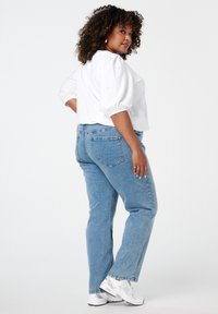 Femme aux cheveux bouclés portant un chemisier blanc orné de perles, un jean bleu clair et des baskets blanches, se tenant avec la main sur la hanche, regardant en arrière.