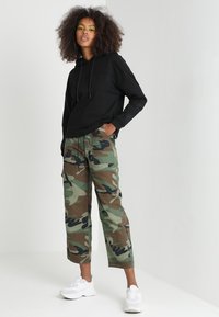 Felpa nera con cappuccio e lacci, pantaloni cargo mimetici in verde e marrone, sneakers bianche. Vestibilità comoda, tasche visibili sui pantaloni.