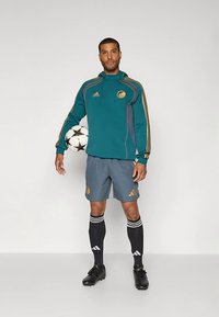 Sweat à capuche de sport bleu sarcelle avec des rayures dorées, short gris, chaussettes de football noires avec des rayures blanches, et chaussures à crampons noires, tenant un ballon de football.