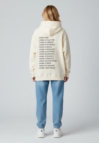 Felpa crema con testo nero che elenca vari interessi, abbinata a pantaloni della tuta azzurri chiari e sneakers bianche. Materiale morbido, vestibilità rilassata.