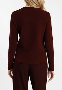 Donna che indossa un maglione a maniche lunghe, testurizzato, di colore marrone scuro e pantaloni coordinati marrone scuro, in piedi con la schiena rivolta verso la telecamera.