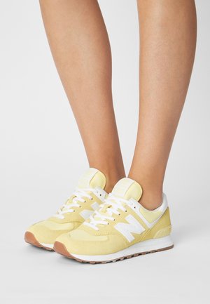 Legs wearing yellow and white New Balance 574 sneakers with white laces, standing on a white surface against a plain background.