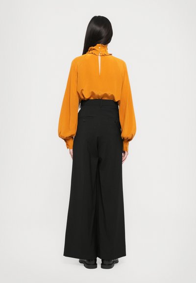 Blusa gialla senape con maniche a sbuffo e colletto arricciato, abbinata a pantaloni neri a gamba larga e scarpe nere. Vista posteriore.
