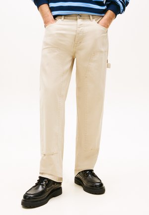 Persona con pantalones beige de pierna recta y bolsillos grandes en los costados, zapatos negros de cuero con cordones, manos en los bolsillos, sobre fondo blanco.