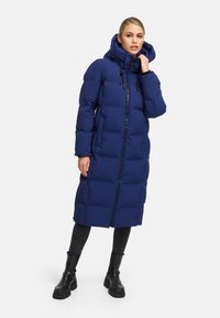 Navy blue puffer coat with a long design, hood, and front zipper. Textured, quilted fabric, with side pockets and a relaxed fit. Black boots.