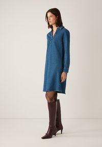 Denim jurk met lange mouwen, kraag en ritsplacket. Draagt gecombineerd met gestreepte maillots en knie hoge bordeaux leren laarzen.