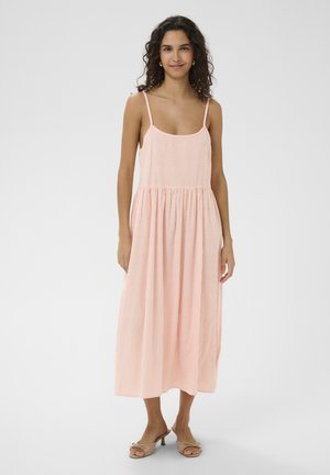 Femme aux cheveux bouclés portant une robe midi sans manches à rayures roses claires et blanches, et des talons ouverts beiges, debout sur un fond blanc.