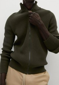 Sudadera de punto con cremallera en verde oliva. Presenta un cuello alto, textura acanalada y puños ajustados. Diseño minimalista con una cremallera suave.
