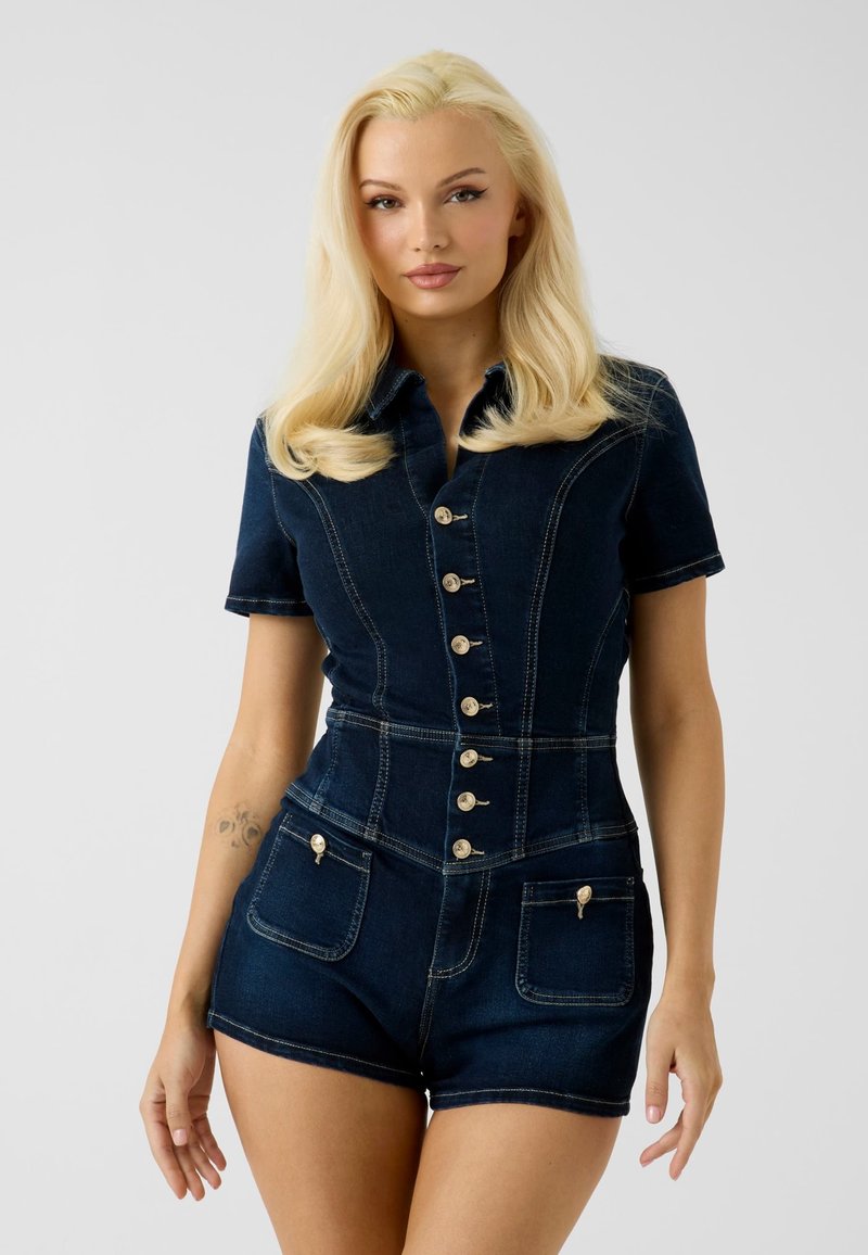 Jeune femme aux longs cheveux blonds portant une combinaison courte en denim bleu foncé à manches courtes boutonnée à l'avant avec des poches, debout devant un fond uni.