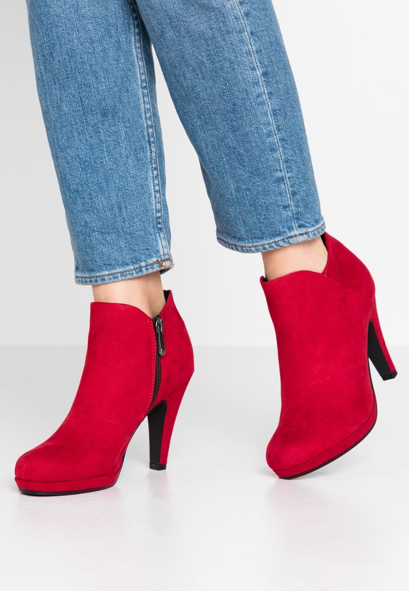 Femme portant un jean bleu et des bottines en daim rouge vif à talons hauts noirs, debout sur une surface blanche.
