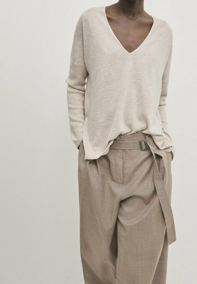 Personne portant un pull beige à col en V, rentré dans un pantalon large taille haute couleur sable avec ceinture en tissu et les mains dans les poches, sur un fond uni.