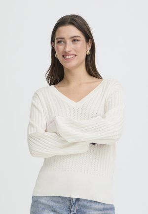 Giovane donna sorridente con le braccia incrociate, indossa un maglione bianco testurizzato a scollo a V e jeans blu su uno sfondo semplice.