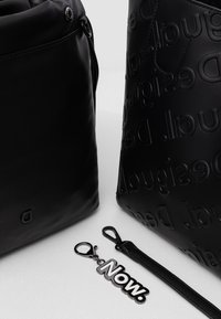 Deux sacs de designer noirs avec des logos en relief, accompagnés d'un porte-clés noir portant le mot « Now. » sur une surface blanche.