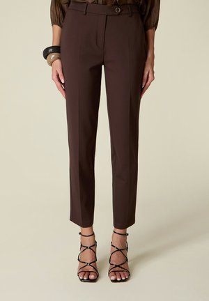 Pantalones ajustados de color marrón oscuro, usados por una persona con un top marrón transparente, sandalias negras de tacón con tiras y pulseras en una muñeca.