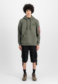 Sudadera verde oliva con un bolsillo delantero y un parche, combinada con pantalones cortos tipo cargo negros y zapatos marrones con estampado. Diseño simple y tejido texturizado.