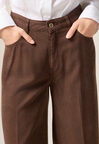 Pantalones anchos marrones hechos de una suave tela de algodón, con bolsillos frontales, un botón de metal y un sutil detalle de costura.