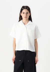 White short-sleeve collared shirt with a textured pattern and a single chest pocket, paired with dark loose-fit pants.