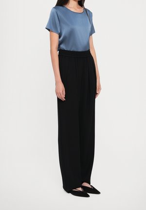 Femme portant un chemisier en satin bleu à manches courtes, un pantalon noir large taille haute et des ballerines noires pointues sur fond blanc.