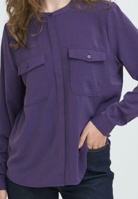 Donna che indossa una camicia a maniche lunghe viola con taschini sul petto abbottonati, mano in tasca, abbinata a jeans blu scuro, tagliati al collo e in vita.