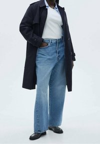 Osoba oblečená v dlouhém tmavém kabátu, bílém tričku, širokých modrých džínách a černých botách, stojící s jednou rukou v kapse.