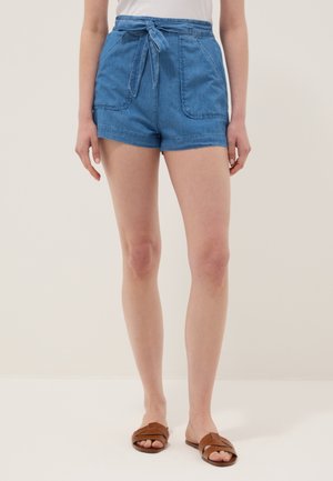 Donna che indossa pantaloncini di jeans blu a vita alta con tasche frontali e cintura annodata, abbinati a sandali marroni con punta aperta.