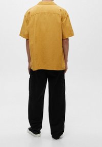 Gelbes kurzärmliges Hemd mit Kragen, das eine subtile Textur aufweist. Kombiniert mit lockeren schwarzen Hosen und weißen Sneakers.