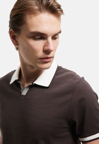 Bruine polo shirt met een witte kraag, met een textuurdetail bij de knopenlijst en ribbelaccenten aan de mouwen. Gladde katoenen stof.
