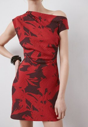 Vestido asimétrico de flores rojas y oscuras sin hombros en una persona de pie con una pulsera negra en la muñeca izquierda sobre un fondo liso.