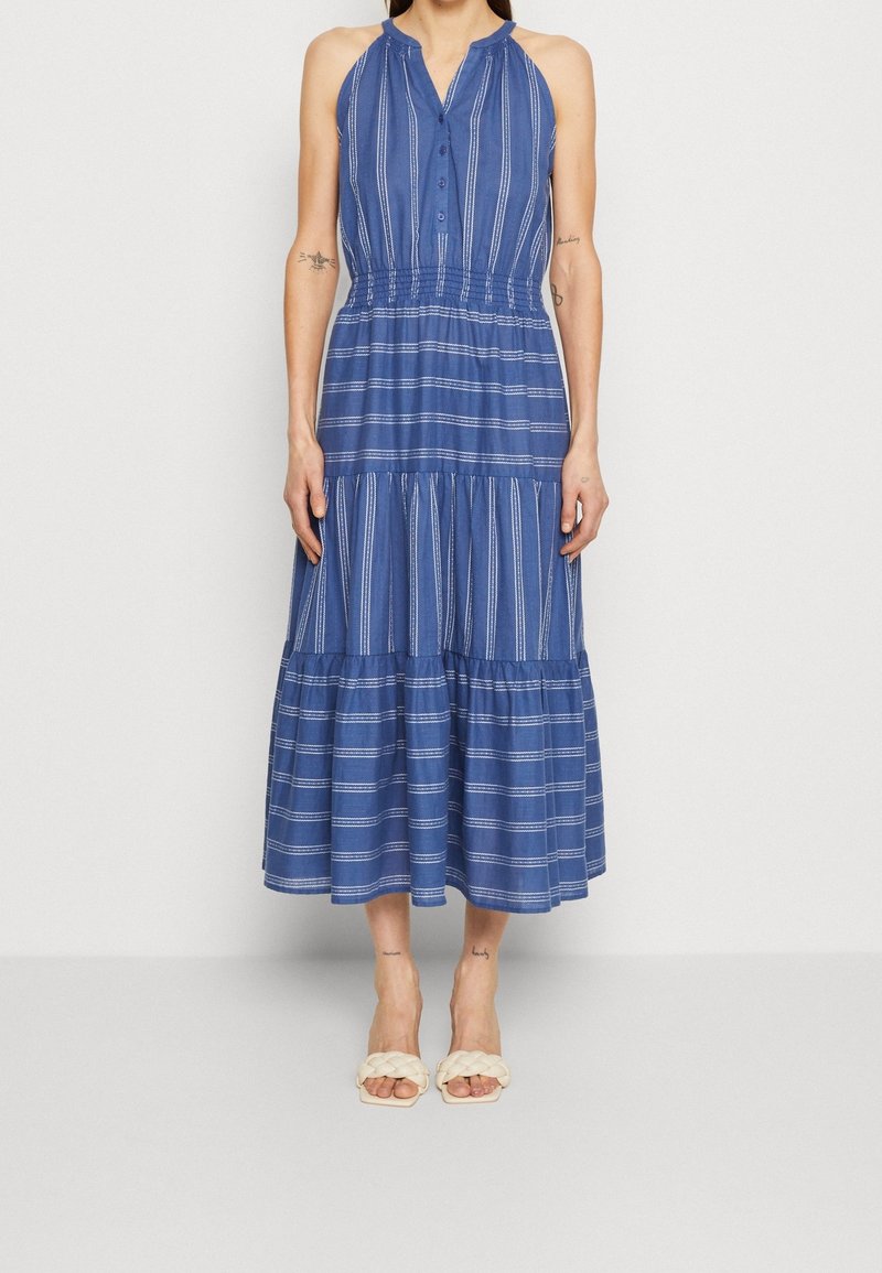 Robe midi bleue sans manches avec des rayures blanches verticales et horizontales, encolure boutonnée, taille élastique, portée avec des sandales blanches tressées à bout ouvert.
