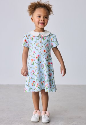 Naeratav noor tüdruk lokkis juustega, kes kannab helehalli lillelist kleiti valge kraega ja valgeid kingi roosade paeltega, seisab hallil põrandal.