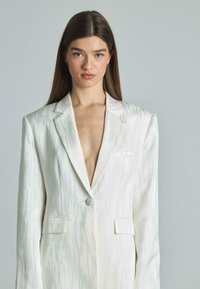 Ivory blazer met een textuur en glans, scherpe revers, enkele knoopsluiting en twee voorzakken. Diepe V-halslijn voegt contrast toe.