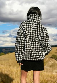 Jachetă din flanel cu model houndstooth alb și negru, cu guler înalt, purtată alături de o fustă neagră, în aer liber, pe un fond de cer înnorat.