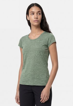 Junge Frau mit langen dunklen Haaren, die ein grünes T-Shirt mit kurzen Ärmeln und schwarze Hosen trägt und leicht nach links schaut.