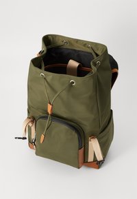 Sac à dos en nylon vert avec une fermeture à cordon, des accents en cuir, une poche zippée à l'avant et des détails de sangle en tissu. L'intérieur semble spacieux et non doublé.