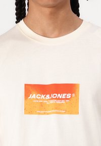 Crème-kleurig T-shirt met een gedrukt oranje rechthoekig logo met witte tekst "JACK&JONES", korte mouwen en een ronde halslijn.