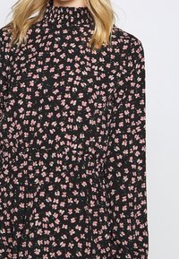 Abito nero floreale con colletto arricciato, maniche lunghe e una fantasia di piccoli fiori rosa e bianchi su uno sfondo scuro.