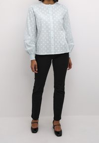 Hellblaue Bluse mit Knopfleiste, strukturiertem Polka-Dot-Muster, gerafften Ballonärmeln und Spitzenakzenten, kombiniert mit schlanken schwarzen Hosen und High Heels.