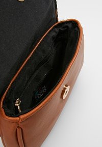 Ruskea nahkalaukku mustalla kankaisella sisäpuolella, jossa näkyy vetoketjullinen sisätasku ja kultainen napitusläpän solki.