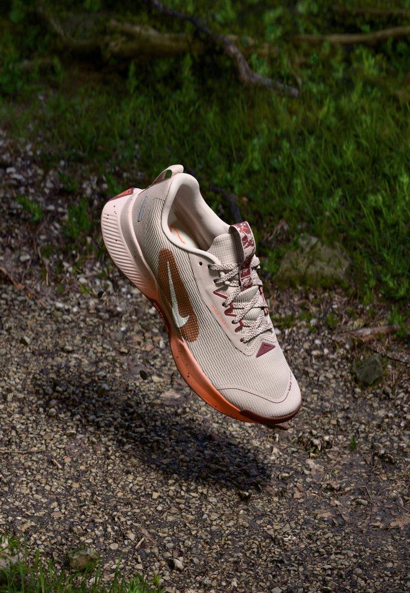 Αθλητικό παπούτσι σε ανοιχτό μπεζ με υφασμάτινο επάνω μέρος με υφή, μπορντό λεπτομέρειες και χαρακτηριστική πορτοκαλί σόλα, σχεδιασμένο για τρέξιμο ή προπόνηση.