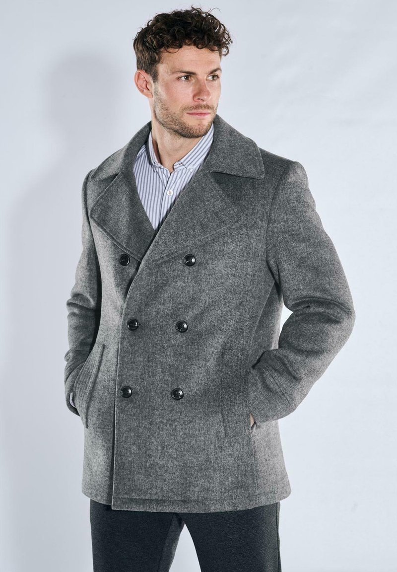 Grau melierter Wollmischmantel mit doppelreihiger Knopfleiste, großen schwarzen Knöpfen, Seitentaschen und breitem Kragen, getragen über einem gestreiften Hemd.