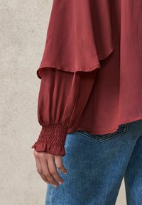 Blouse rouge à manches longues avec un design superposé et des poignets smockés, associée à un jean en denim bleu. Tissu doux avec une texture fluide.