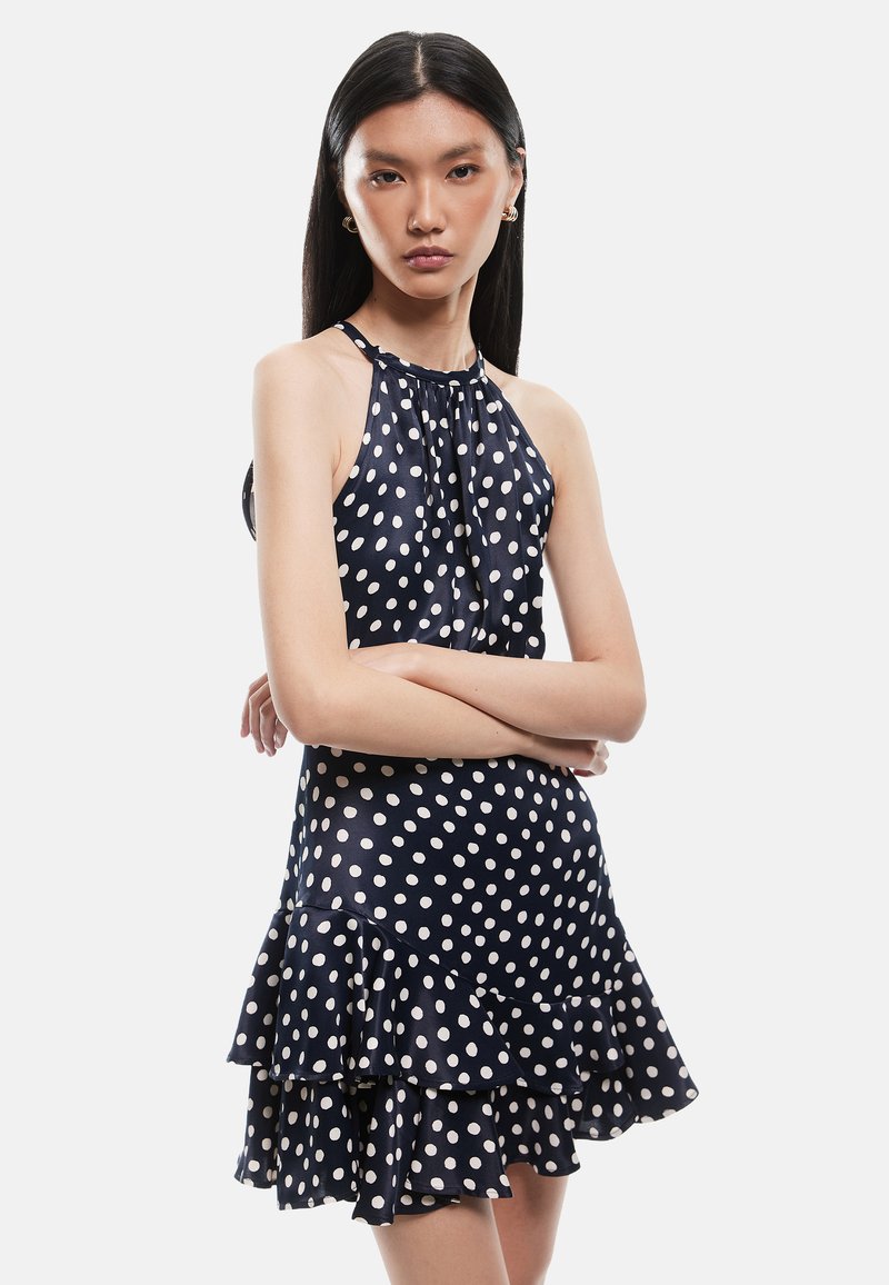 Vestido de halter náutico con lunares blancos, que presenta un dobladillo con volantes y una tela ligera. El diseño incluye un escote recogido y una silueta ajustada.