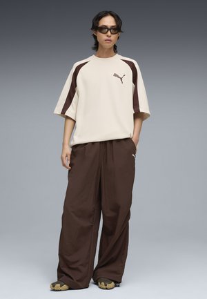 Persona che indossa abbigliamento sportivo Puma beige e marrone oversize, occhiali da sole scuri e scarpe marroni massicce, in piedi davanti a uno sfondo grigio.