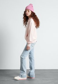 Ljusrosa hoodie med ficka fram, kombinerad med ljusblå bootcut-jeans. Modellen bär en rosa mössa och vita sneakers med röda detaljer.