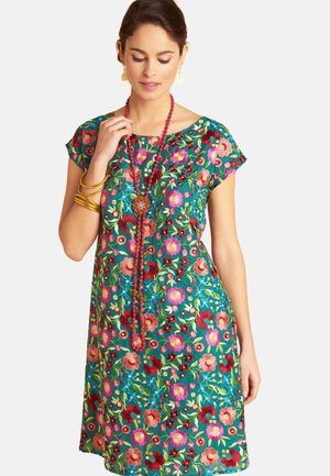 Woman wearing short-sleeve teal dress with colorful floral embroidery, red beaded necklace, gold bangles, and earrings, looking down.