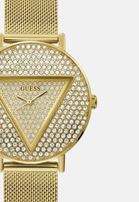Guld mesh rem ur med rund glitrende urskive. Trekantmønster af funkende rhinsten omkranser urhåndledene og mærkenavnet.